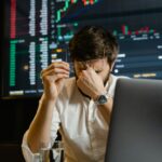 A stressed man looks at stock market data on his computer screen in an office setting.
